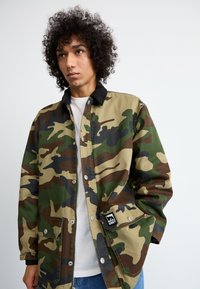 Καμουφλάζ τζάκετ με πράσινα, καφέ και μαύρα σχέδια, ανθεκτικό ύφασμα, κουμπιά με κλιπ και μαύρη ετικέτα με λογότυπο στην αριστερή τσέπη.