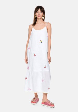 Frau trägt ein weißes ärmelloses Midikleid mit bunten gestickten Motiven und rosa glitzernden Sandalen mit Schnallen, stehend vor einem einfarbigen Hintergrund.