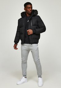 Schwarze Pufferjacke mit einem mit Fell gefütterten Kapuze, Reißverschluss- und Knopfverschlüssen, getragen über einem schwarzen Shirt und grauen Jogginghosen, kombiniert mit weißen Sneakers.