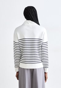 Maglione bianco a righe con strisce orizzontali blu navy, con colletto morbido, polsini e orlo a coste. Indossato su pantaloni a gamba larga grigio chiaro.