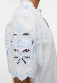 Blusa bianca con maniche corte a sbuffo, caratterizzata da ricami blu e motivi floreali intagliati lungo le maniche e l'orlo.
