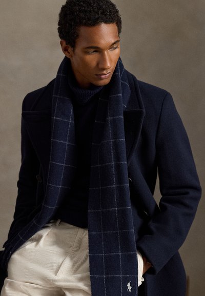 Navy vuneni kaput dvostrukog kopčanja, teksturirani mornarski pamučni rolka džemper i karirani šal, uparen s bež manšesternim hlačama.
