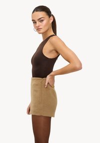 Bodysuit sans manches marron associé à une mini-jupe en fausse fourrure beige avec une fermeture éclair à l'arrière et un détail doré rond.