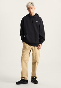 Felpa nera con cappuccio e tasca frontale, pantaloni beige e sneakers nere con logo bianco. Capelli corti, texturizzati e di colore chiaro.