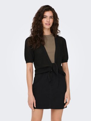 Femme portant un cardigan en maille noir à manches courtes noué à la taille, un haut vert olive et une jupe noire, debout devant un fond clair uni.