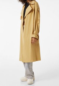 Longue trench beige avec un large col, des poignets boutonnés et une coupe droite, portée sur un haut noir et un jean gris clair. Chaussures blanches épaisses.