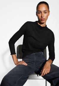 Schwarzes, geripptes Langarmoberteil mit einem gerafften Ausschnitt, kombiniert mit hochgeschnittenen dunklen Jeans. Die Textur ist glatt und figurbetont.