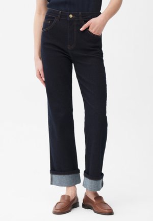 Persoon draagt donkerblauwe opgerolde jeans met bruine loafers en een zwart geribd shirt, staat met één hand in de zak.