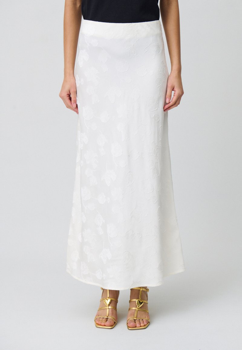 Falda maxi blanca con textura floral, presenta un acabado suave y una cintura ajustada. Combinada con sandalias doradas de tiras.