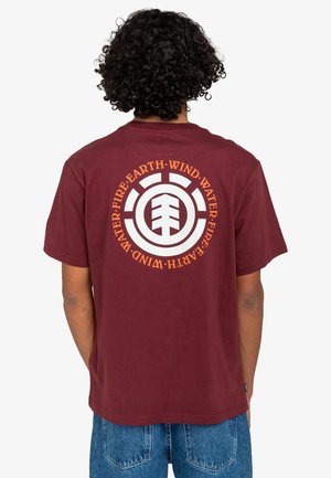 Person med krøllet hår iført en vinrød t-shirt med et cirkulært logo og teksten "Ild Jord Vind Vand" på ryggen, kombineret med blå jeans.