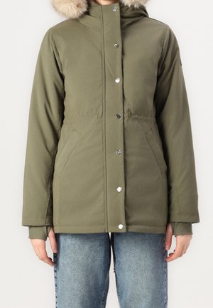 Zielona oliwkowa zimowa kurtka z kapturem, obszyta futrem i zapinana na srebrne zatrzaski, noszona z jasnoniebieskimi dżinsami.