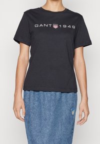 En person iført en sort GANT 1949 t-shirt og en blå nederdel med et lille trekantet logo-patch på højre side.