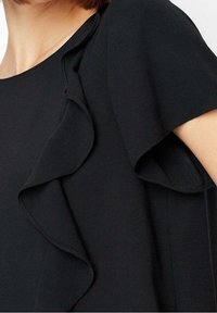 Haut noir avec un col rond et des manches courtes, avec des volants superposés sur le devant, fabriqué dans un tissu lisse.