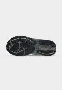 Semelle d'une basket New Balance noire et grise montrant des motifs de grip texturés et le nom de la marque au niveau du talon.