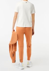 Person som bär en vit kortärmad skjorta och orange joggers, håller en matchande orange jacka, står med vita sneakers på vit bakgrund.