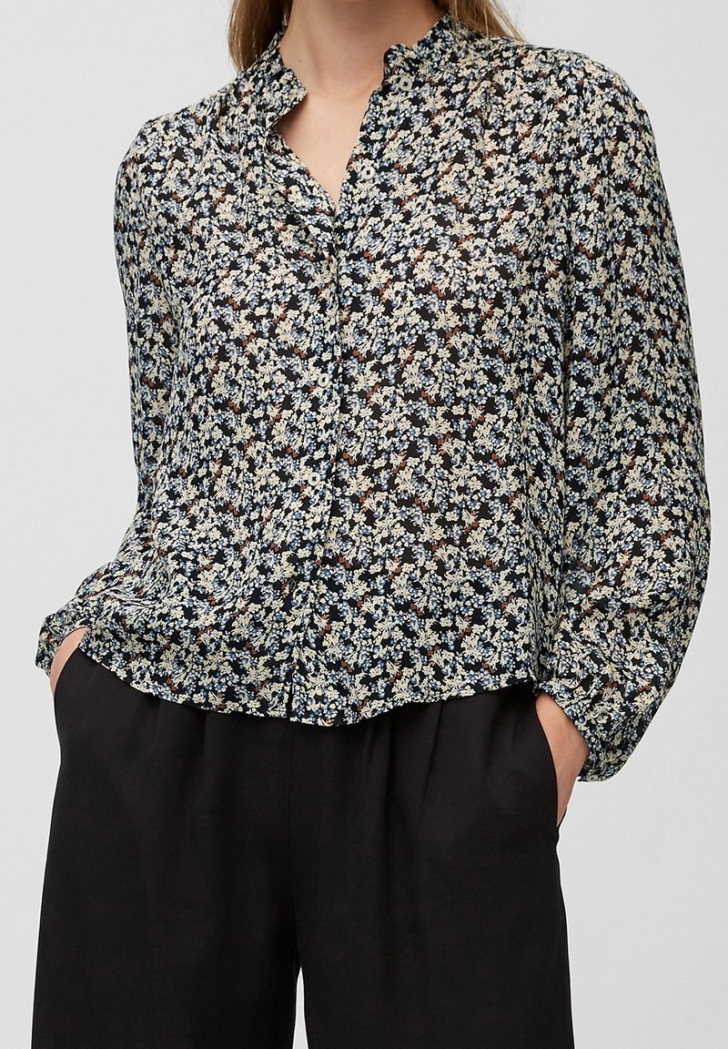 Femme portant un chemisier à manches longues à boutons avec un petit imprimé floral et un pantalon noir, les mains dans les poches.