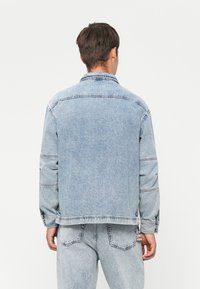 Man facing away wearing light blue denim jacket and matching jeans against plain white background.