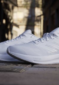 Paar weiße Sportschuhe mit Schnürsenkeln und Cloudfoam-Sohlen, platziert auf einem Steinpflaster in einem städtischen Außenbereich.