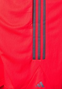 Tissu rouge avec trois rayures noires verticales et un petit logo Adidas gris dans le coin inférieur droit.