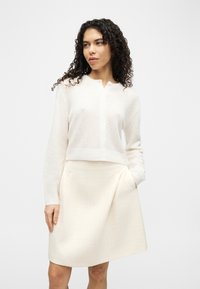 Γυναίκα με σγουρά μαλλιά που φοράει λευκή ζακέτα με κουμπιά και υφασμάτινη φούστα σε σχήμα A, στέκεται με το ένα χέρι στην τσέπη, μπροστά σε απλό φόντο.