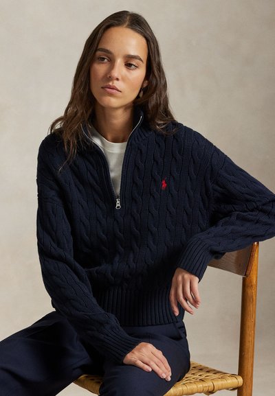 Joven sentada en una silla de madera, lleva un suéter azul marino con trenzado y cremallera media y pantalones oscuros, mirando ligeramente a la izquierda.