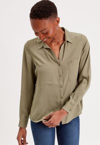 Chemise olive, à manches longues et boutons, avec deux poches poitrine, en tissu doux, et une coupe décontractée avec col et poignets à boutons.