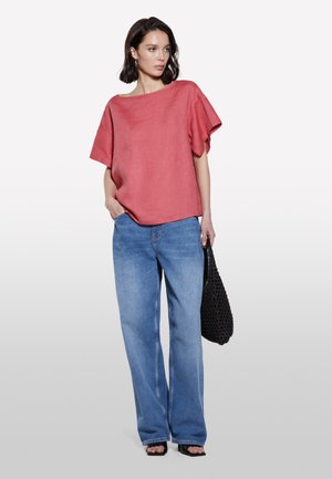 Haut rouge en lin à manches courtes associé à un jean en denim bleu clair à jambes larges. Le modèle tient un sac noir texturé et porte des sandales plates.