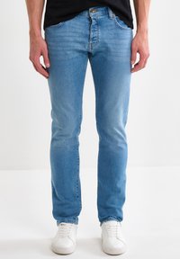 Hombre con jeans ajustados azul claro, camisa negra y zapatillas blancas, de pie frente a un fondo blanco liso.