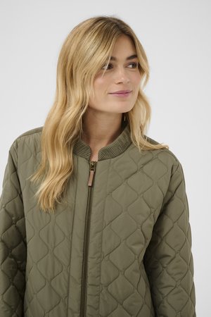 Pitkät vaaleat hiukset omaava nainen pukeutuneena oliivinvihreään tikattuun takkiin, jossa on resorikaulus, katse suunnattu hieman oikealle.