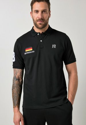 Mężczyzna z brodą ubrany w czarną koszulkę polo z naszywką niemieckiej flagi, napisem "Deutschland" oraz białym logo "J PI π", stoi z ręką w kieszeni.