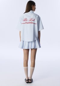 Light blue striped short-sleeve shirt with a curved hem, featuring red embroidered text on the back. Accompanied by a pleated, matching skirt.