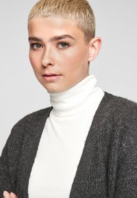 Cardigan en tricot gris avec front ouvert, en tissu texturé, porté sur un haut à col roulé blanc. Le modèle avec les cheveux blonds courts fait face à la caméra.
