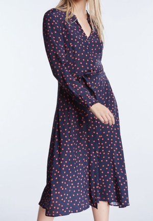 Tmavě modré šaty z lehké látky s dlouhými rukávy, s červeným puntíkovaným vzorem, zapínáním na knoflíky vpředu a páskem v pase ve stejném stylu.