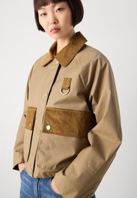 Beige jasje met een bruin corduroy kraag en details. Voorzien van voorzakken, drukknoopsluiting en een metalen lusaccent.