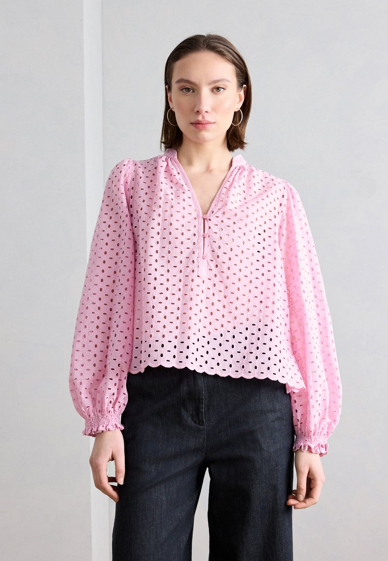 Naine, kes kannab heleroosat pika varrukaga õhulist pluusi sakilise äärega ja mustaid pükse, seisab ühevärvilise halli tausta ees.