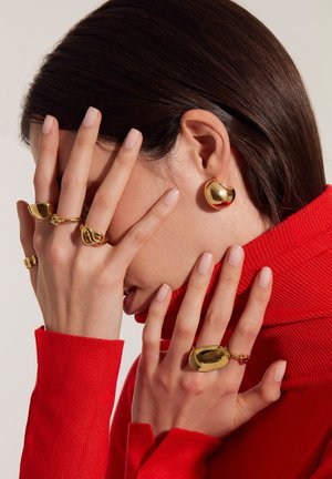 Gouden oorbellen en ringen tentoongesteld op handen, met gladde, ronde vormen. Het model draagt een aansluitende rode coltrui. Zacht licht benadrukt de texturen.
