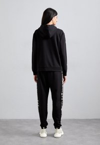 Haut à capuche noir avec une poche kangourou à l'avant et un cordon de serrage, associé à un pantalon de jogging assorti avec des accents de logo en or.