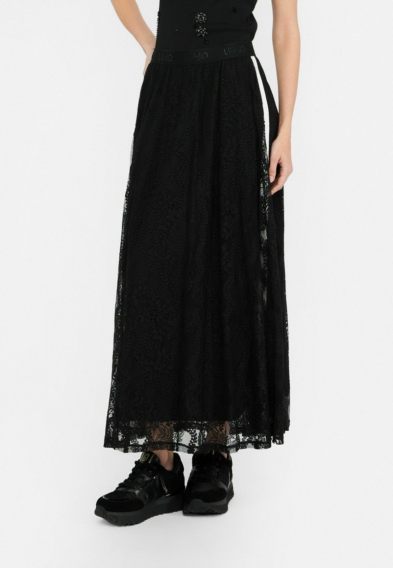 Schwarzer Maxi-Rock aus Spitze mit einem strukturierten Muster und elastischem Bund, getragen über schwarzen Sneakers mit glänzenden Akzenten.