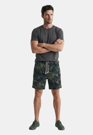 Hombre con los brazos cruzados vistiendo una camiseta gris oscura, pantalones cortos de camuflaje con cordones blancos y zapatillas verdes, de pie frente a un fondo liso.
