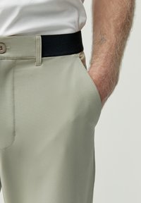 Beiga byxor med en slät textur, med ett svart elastiskt midjeband och en sidficka. Knäppning i midjan. Nära bild.