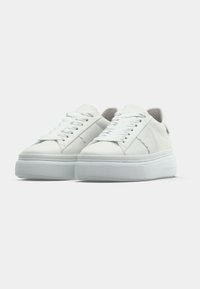 Λευκό αθλητικό παπούτσι με επάνω μέρος από λείο δέρμα, επίπεδα κορδόν ια και παχύ, υφασμένο ελαστικό πάτο πλατφόρμας. Διαθέτει ελάχιστες ραφές και διακριτικές λεπτομέρειες με λογότυπο.