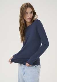 Navy long-sleeve shirt in soft fabric, scoop neckline, slim fit, paired with light blue denim jeans. Smooth texture, minimal design.
