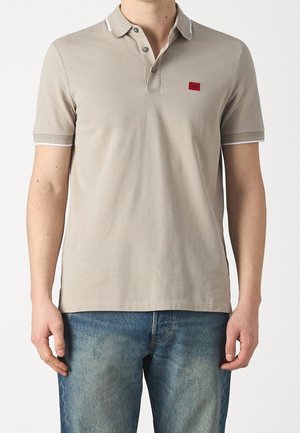 Mann trägt hellbeiges kurzärmeliges Polohemd mit einem roten quadratischen Logo auf der Brust und blaue Jeans aus Denim, steht vor weißem Hintergrund.