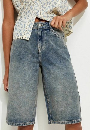 Kobieta w wysokich, luźnych, sięgających do kolan szortach dżinsowych i bezrękawowej, kwiatowej bluzce z koronkowym wykończeniem, ręką poprawiająca pas spodni.