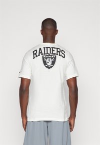 Camiseta de algodón blanca con un gran texto y logo en negro de "RAIDERS" en la parte posterior. Manga corta y cuello redondo.