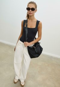 Body nero senza maniche, pantaloni bianchi a gamba larga, borsa nera e sneakers marroni chiare. Modella con occhiali da sole scuri su uno sfondo neutro.