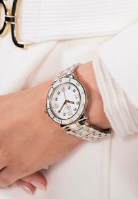 Srebrnast i zlatan ručni sat s okruglim bijelim brojčanikom, rimskim brojevima, oznakama koje nalikuju na dijamante i metalnom narukvicom.