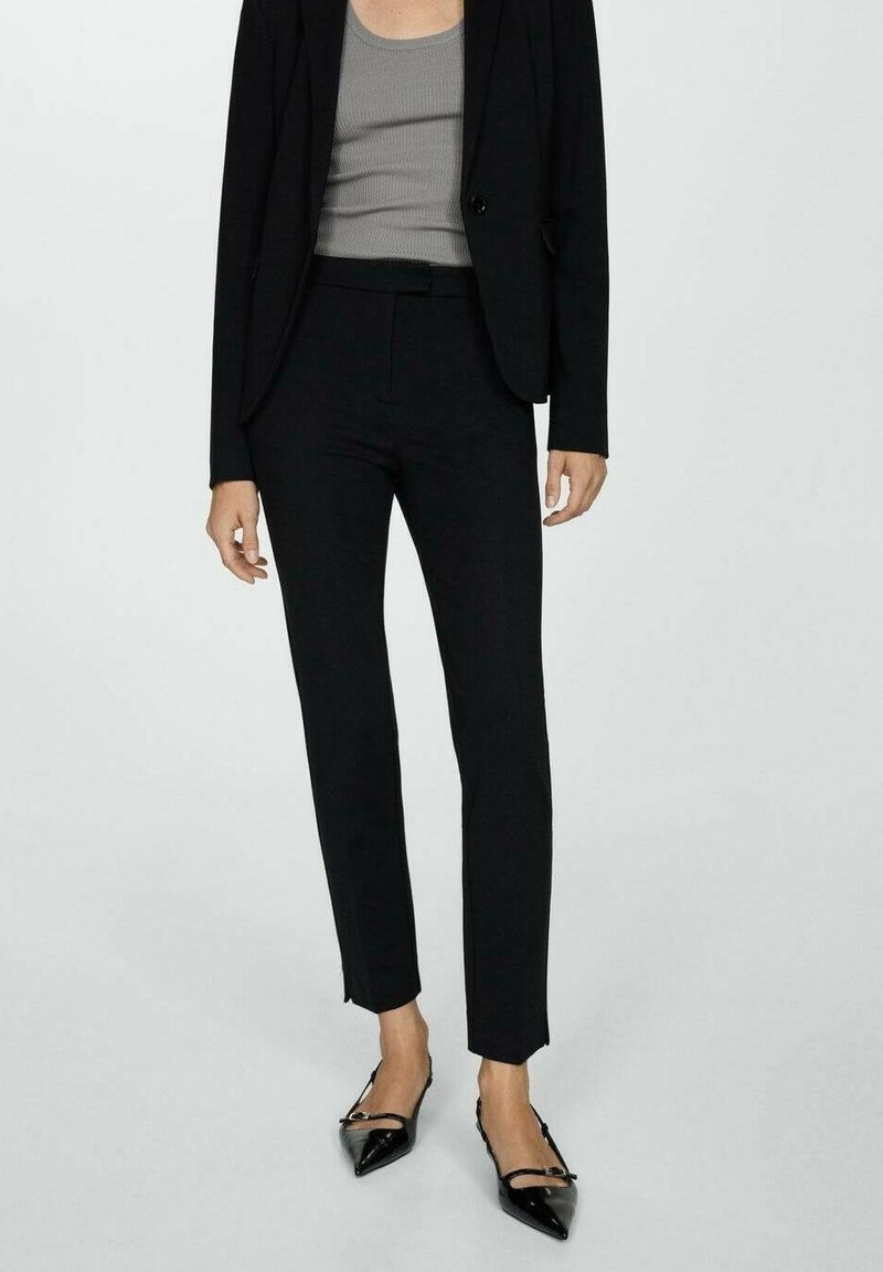 Veste de costume noire ajustée et pantalon avec un débardeur gris. Le pantalon présente une coupe slim et un style minimaliste. Chaussures noires à bouts pointus.