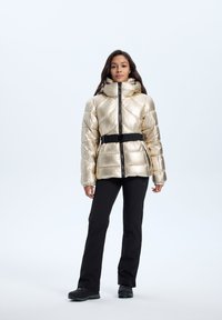 Metallisk gylden pufferjakke med høj krave, indsnævret talje med et sort bælte, lynlås foran og struktureret quiltet mønster. Sorte bukser og sko.