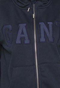 Sudadera con cremallera color navy, hecha de una tela suave, con grandes letras bordadas en azul oscuro que dicen "GANT", cremallera plateada y cuello con cordón.